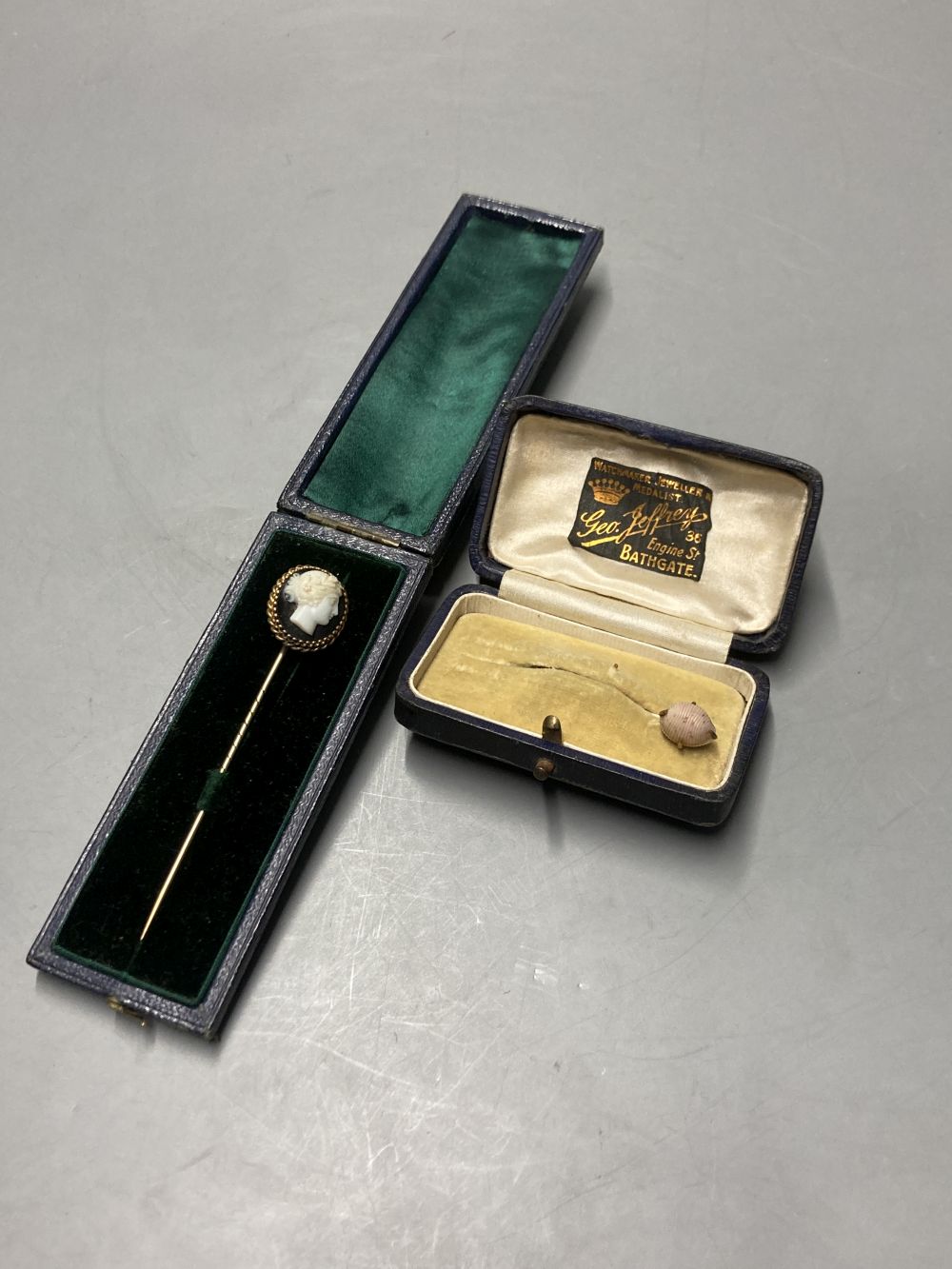 A cased early 20th century yellow metal and carved sardonyx set stick pin, 8cm and a similar shell stick pin, 56mm.